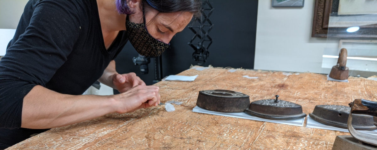 Photo of a pale olive woman mending the reverse side of a large tapa bark cloth. She is wearing a black shirt and black and gold face mask and is placing a pasted piece of kozo mulberry paper with a pair of tweezers. Across the back cloth previous mends are drying under blotter paper and weights.